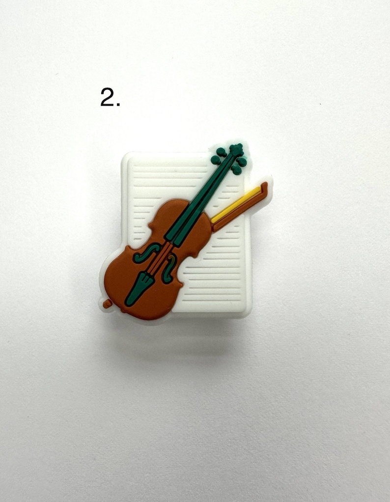 May include: A white charm with a brown violin on top of a sheet of music. The violin is green and yellow.
