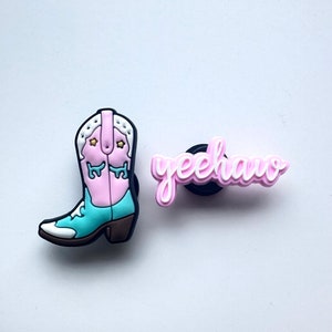 May include: Two pink and white shoe charms. One charm is a cowboy boot with a turquoise top and gold stars. The other charm says "yeehaw" in white letters.