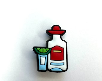 May include: A cartoon illustration of a tequila bottle with a sombrero and a shot glass with a lime wedge. The bottle is white with a red label and a black outline. The shot glass is blue with a white interior and a green lime wedge. The sombrero is red with a black outline.