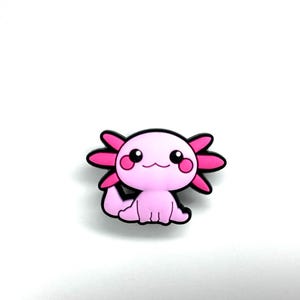 Puede incluir: Un dije en forma de ajolote rosa con contornos negros. El dije presenta una cara sonriente, ojos negros grandes y branquias rosas. El ajolote está sentado sobre un fondo blanco.