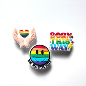 Può includere: Tre spille smaltate con colori arcobaleno e design diversi. La prima spilla presenta un paio di mani che tengono un cuore arcobaleno. La seconda spilla presenta un viso sorridente arcobaleno con il testo "Be Fabulous" sotto. La terza spilla presenta il testo "Born This Way" in colori arcobaleno.