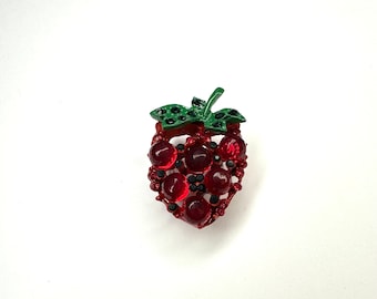 May include: A red rhinestone strawberry brooch with a green leaf. The strawberry is made of metal and has a shiny finish.
