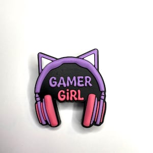 May include: A purple and black charm featuring a headset design with cat ears. The text "GAMER GIRL" is displayed in pink and purple. The charm is likely made of rubber or a similar material.