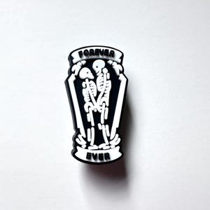 May include: Black and white enamel pin featuring two skeletons holding each other in a coffin. The pin says "Forever Ever" on a banner.
