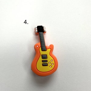 May include: A small orange and yellow plastic guitar charm with black details. The guitar has a black pickguard and white knobs.