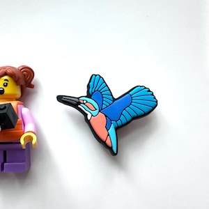 May include: A colorful shoe charm featuring a stylized blue and orange bird with outstretched wings. The bird has a black beak and is set against a white background. The charm is likely made of rubber or a similar material.