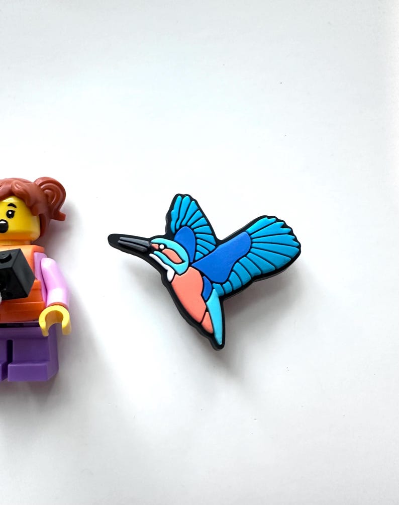 May include: A colorful shoe charm featuring a stylized blue and orange bird with outstretched wings. The bird has a black beak and is set against a white background. The charm is likely made of rubber or a similar material.