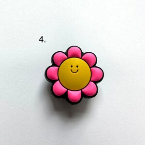 May include: A pink flower charm with a yellow smiley face in the center. The charm is made of rubber and has a glossy finish.