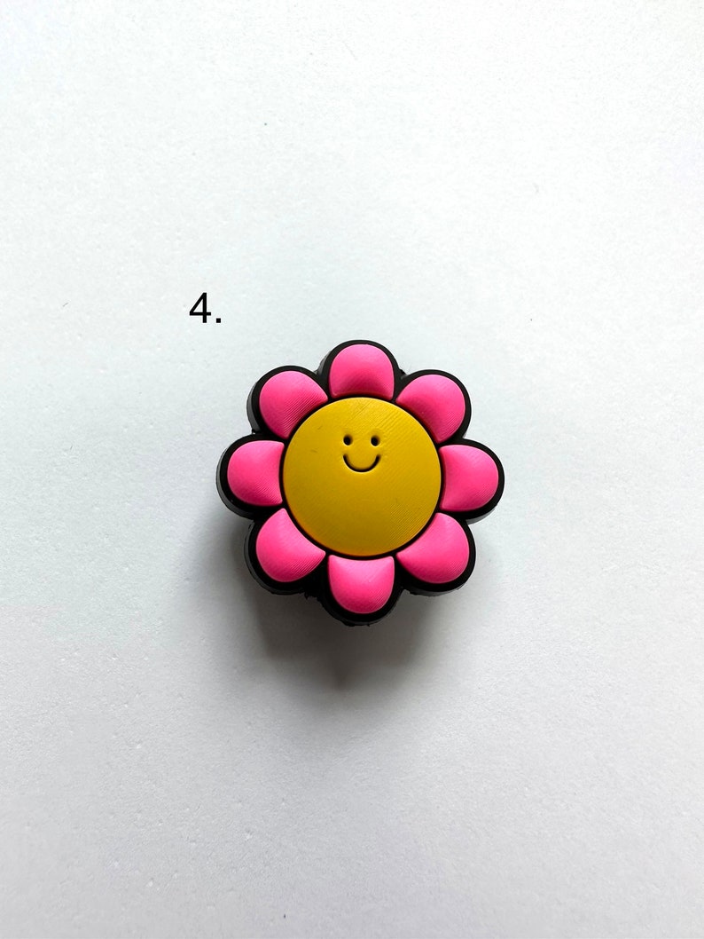 May include: A pink flower charm with a yellow smiley face in the center. The charm is made of rubber and has a glossy finish.
