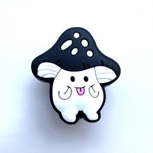 May include: A black and white mushroom character with a pink tongue sticking out. The mushroom has white spots on its cap and a white body with arms and legs.