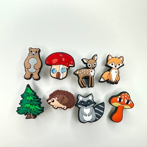 May include: A set of nine colorful cartoon animal and nature charms, including a bear, a mushroom house, a deer, a fox, a pine tree, a hedgehog, a raccoon, and a mushroom. The charms are made of rubber and are perfect for adding a whimsical touch to any project.
