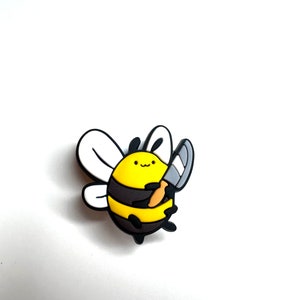 May include: A yellow and black bee-shaped charm with a smiling face and a silver knife. The bee has white wings and is set against a white background. The charm is likely made of rubber or a similar material.