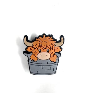 May include: A brown and white Highland cow cartoon charm with a fluffy mane and horns, sitting in a gray wooden bucket.