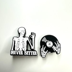 May include: Two black and white enamel pins. One pin features a skeleton giving a thumbs up with the text "NEVER BETTER". The other pin features a skeleton hand holding a black vinyl record.