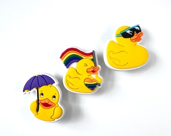 Könnte beinhalten: Drei gelbe Gummi-Enten-Anhänger mit weißen Rändern. Eine Ente hält einen lila Regenschirm, eine andere eine Regenbogenflagge und ein Herz, und die dritte trägt eine Sonnenbrille. Alle drei vor weißem Hintergrund.