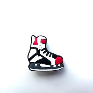 May include: A white and black hockey skate charm with red accents. The skate is made of a rubber-like material and is designed to look like a traditional hockey skate.