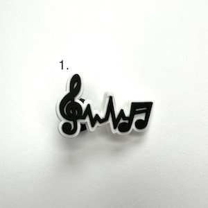May include: Black and white charm featuring a treble clef, a heartbeat, and a musical note. The charm is made of rubber and is designed to be attached to a shoe or other item.