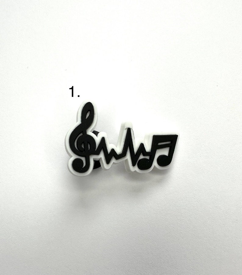 May include: Black and white charm featuring a treble clef, a heartbeat, and a musical note. The charm is made of rubber and is designed to be attached to a shoe or other item.