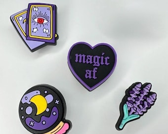 Op de afbeelding: Een set van vier schoenbedels met een paarse tarotkaart met een oog, een zwart hartvormig bedeltje met de tekst magic af in het paars, een ronde kristallen bol met een gele maan en een takje paarse lavendel met een lichtblauwe steel.