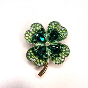 Può includere: Una spilla a forma di quadrifoglio con stelo e cornice color oro. Il quadrifoglio è tempestato di numerose piccole pietre rotonde verde chiaro e iridescenti, arricchite da quattro grandi pietre sfaccettate color verde smeraldo a goccia.