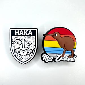 Może przedstawiać: Dwie przypinki o tematyce nowozelandzkiej. Jedna to czarno-biała tarcza ze słowem "HAKA" i maoryskim wzorem. Druga to brązowy ptak kiwi na tle zachodu słońca z napisem "New Zealand".