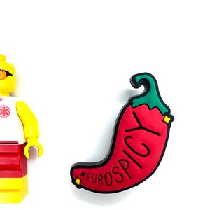 Puede incluir: Un dije en forma de chile rojo con la palabra "NEUROSPICY" en letras negras. El dije tiene un tallo verde y detalles amarillos. Una figura de Lego amarilla y roja es parcialmente visible a la izquierda.