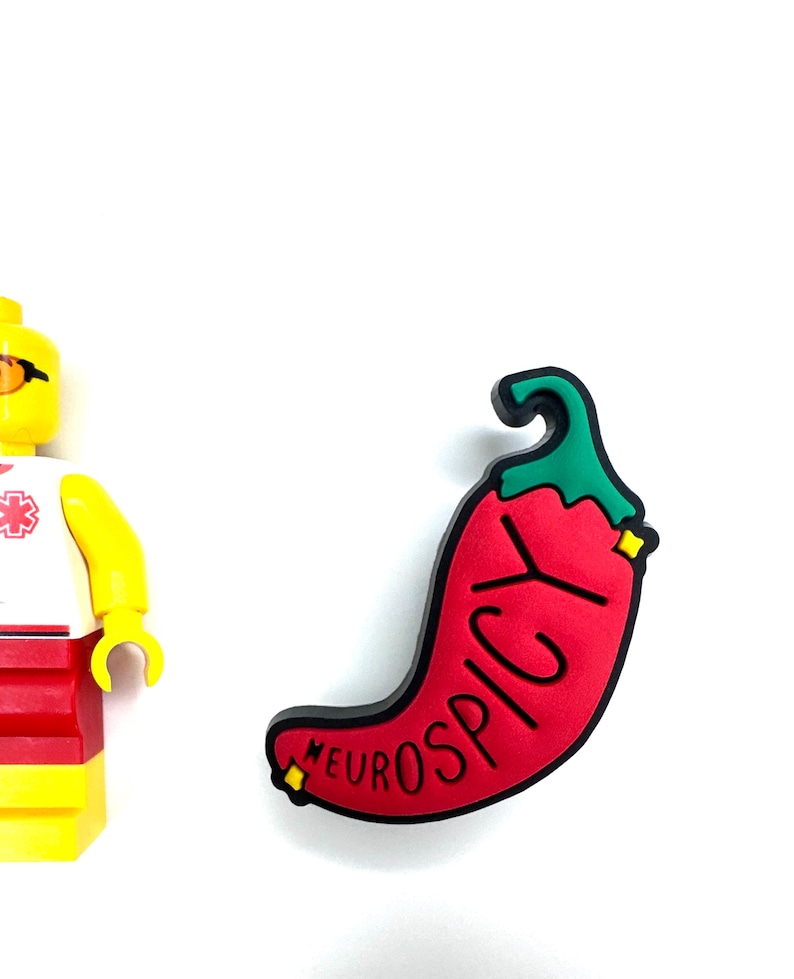 Puede incluir: Un dije en forma de chile rojo con la palabra "NEUROSPICY" en letras negras. El dije tiene un tallo verde y detalles amarillos. Una figura de Lego amarilla y roja es parcialmente visible a la izquierda.
