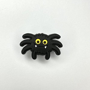 May include: A black spider-shaped charm with yellow eyes and two small white fangs. The spider has eight legs and a textured body. This Halloween-themed charm is suitable for various crafting projects.