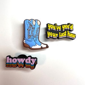 Op de afbeelding: Drie kleurrijke cowboy-thema schoenbedels. Een bedel heeft een paar blauwe cowboylaarzen met witte stiksels. Een andere bedel zegt "howdy" in regenboogkleuren. De derde bedel zegt "you've yee'd your last haw" in gele letters op een zwarte achtergrond.