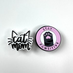 Peut inclure: Deux breloques blanches et noires sur le thème du chat. Un charme dit "cat mom" et l'autre charme dit "Stay Pawsitive" avec une empreinte de patte.