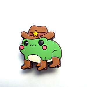 Puede incluir: Una rana verde con un sombrero de vaquero marrón con una estrella amarilla y botas de vaquero marrones. La rana tiene mejillas rosadas y una barriga blanca. La rana es un diseño de estilo caricatura.