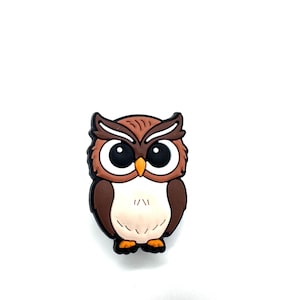 May include: A brown and white owl-shaped charm with large black eyes, a yellow beak, and orange feet. The owl has a black outline and is set against a white background. This charm is suitable for jewelry or craft projects.