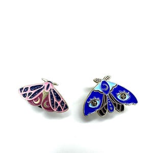 May include: Two moth-shaped enamel pins. One is pink, navy, and burgundy, the other is blue, black, and light blue. Both have detailed wing patterns and celestial designs.
