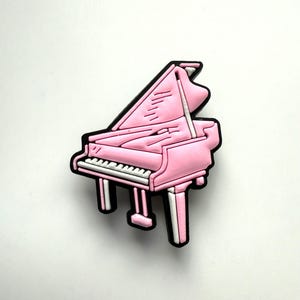 Könnte beinhalten: Ein rosa Flügel-Charm mit schwarzer Umrandung und weißen Tasten. Das Klavier hat eine glänzende Oberfläche und ist vor einem weißen Hintergrund platziert. Der Charm ist eine Miniaturdarstellung eines Musikinstruments.