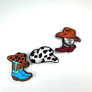 Op de afbeelding: Drie cowboy laarzen en hoed charms. Een charm is een bruine cowboyhoed met een rode laars. Een andere charm is een zwart-witte koe-print cowboyhoed. De laatste charm is een bruine en oranje luipaardprint cowboyhoed met een blauwe laars.