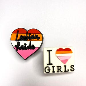 Op de afbeelding: Twee schoenbedels. De ene is een hartvormige bedel met horizontale strepen in rood, oranje, wit en roze, met de zwarte tekst "Lesbian Pride". De andere is een witte rechthoekige bedel met de zwarte tekst "I" en "GIRLS" en een klein hartje.