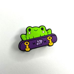 May include: A green frog with a cute face is riding a purple skateboard with yellow wheels. The skateboard has a heart-shaped design on the top.