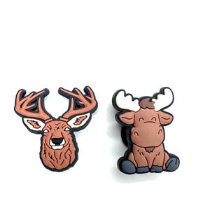 May include: Two animal-themed shoe charms. One is a brown deer head with antlers, and the other is a brown moose with white antlers. Both charms have black outlines and are set against a white background.