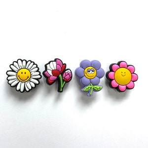 May include: Four colorful flower shoe charms with smiling faces. The charms are made of rubber and feature a daisy, a pink flower, a purple flower, and a yellow flower.