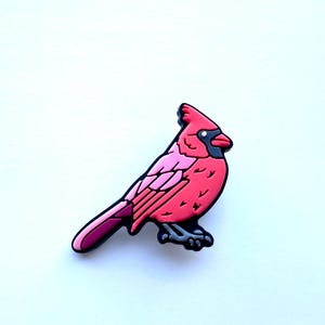 May include: A red cardinal bird charm with a pink belly and black eyes. The charm is made of a rubber-like material.