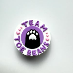 May include: A round, white and purple charm with the words "TEAM TOE BEANS" in purple lettering. The center features a black cat paw print with white toe beans, surrounded by a purple circle and small pink hearts.