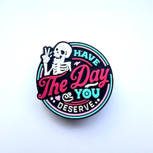 May include: A black and white skeleton with a peace sign, in a pink and blue circle with the text "Have the Day You Deserve".
