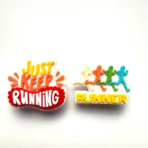 May include: Two vibrant shoe charms with running motifs. One features the text "Just Keep Running" in white on a red and orange design. The other displays four running figures in yellow, orange, green, and blue, with the word "RUNNER" in yellow.