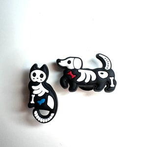Pode incluir: Dois encantos de animais esqueleto em preto e branco. Os encantos são um gato e um dachshund. Ambos os encantos têm um osso branco nas suas patas e o texto "I" nos seus corpos.