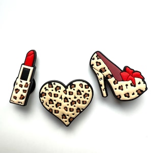 May include: Three decorative charms: a lipstick, a heart, and a high-heeled shoe. Each charm features a tan and brown leopard print pattern. The lipstick has a red top, the heart is heart-shaped, and the shoe has a red bow.