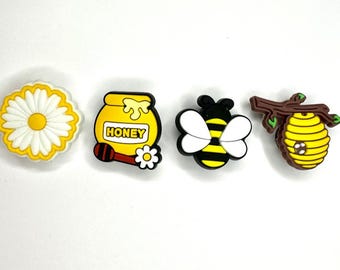 May include: Four yellow and brown bee-themed shoe charms. The charms include a honey pot with the word "honey", a bee, a daisy, and a beehive on a branch.