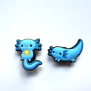 Może przedstawiać: Dwa niebieskie charmsy w kształcie aksolotla z uroczym wzorem kreskówkowym. Jeden aksolotl trzyma żółtą gwiazdę, a drugi pływa.
