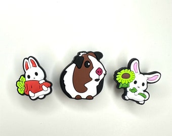 May include: Three white cartoon animal charms with black outlines. A rabbit holding a carrot, a guinea pig, and a rabbit holding a sunflower.