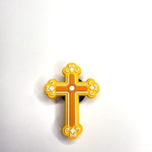May include: A yellow and orange cross-shaped charm with decorative white accents. The cross has a central vertical line and horizontal lines, with a small white circle at the center. The edges of the cross are rounded with decorative swirls.