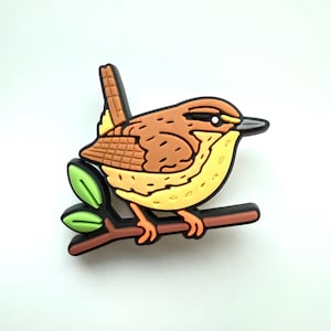 May include: A colourful bird-shaped pin. The bird is perched on a brown branch with green leaves. The bird has brown, yellow, and orange feathers, a black beak, and orange feet. The pin is made of a glossy material.